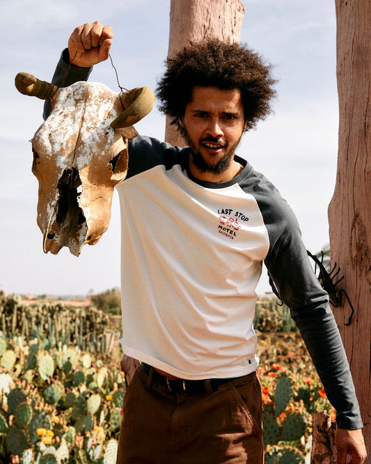 A man with curly hair stands outdoors holding a cattle skull, wearing a Saltrock Last Stop Long Sleeve Raglan in white and brown pants, with cacti in the background.
