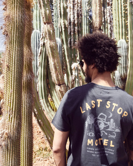 A person with curly hair and sunglasses is standing near tall cactus plants, wearing a dark grey shirt that says "Last Stop Motel" on the back, showcasing Saltrock branding in 100% cotton.