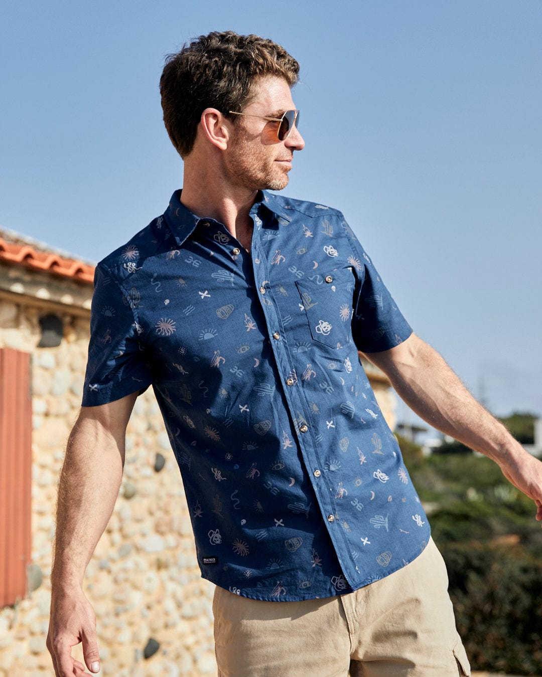 A man wearing a Saltrock Last Stop Mens Short Sleeve Shirt in Blue and khaki shorts.