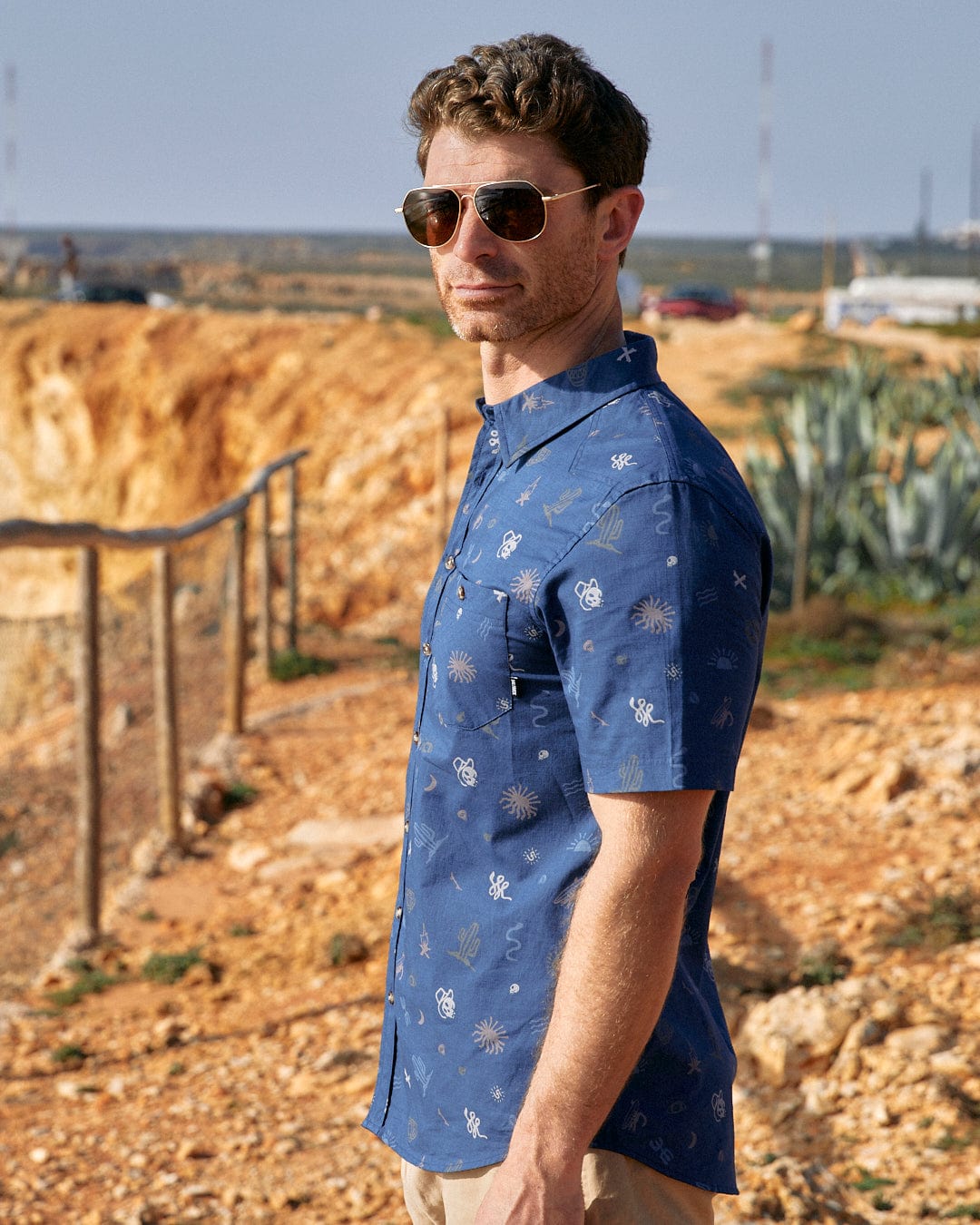 A man in a Last Stop - Mens Short Sleeve Shirt - Blue by Saltrock standing on a dirt road.
