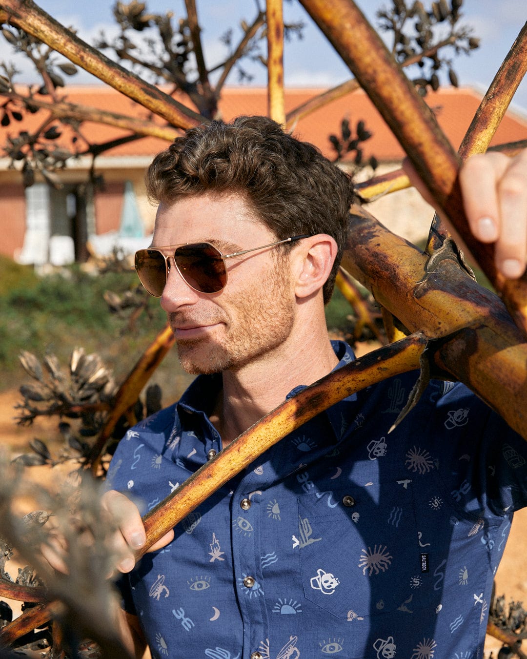 A man in a Last Stop - Mens Short Sleeve Shirt - Blue by Saltrock leaning against a tree.