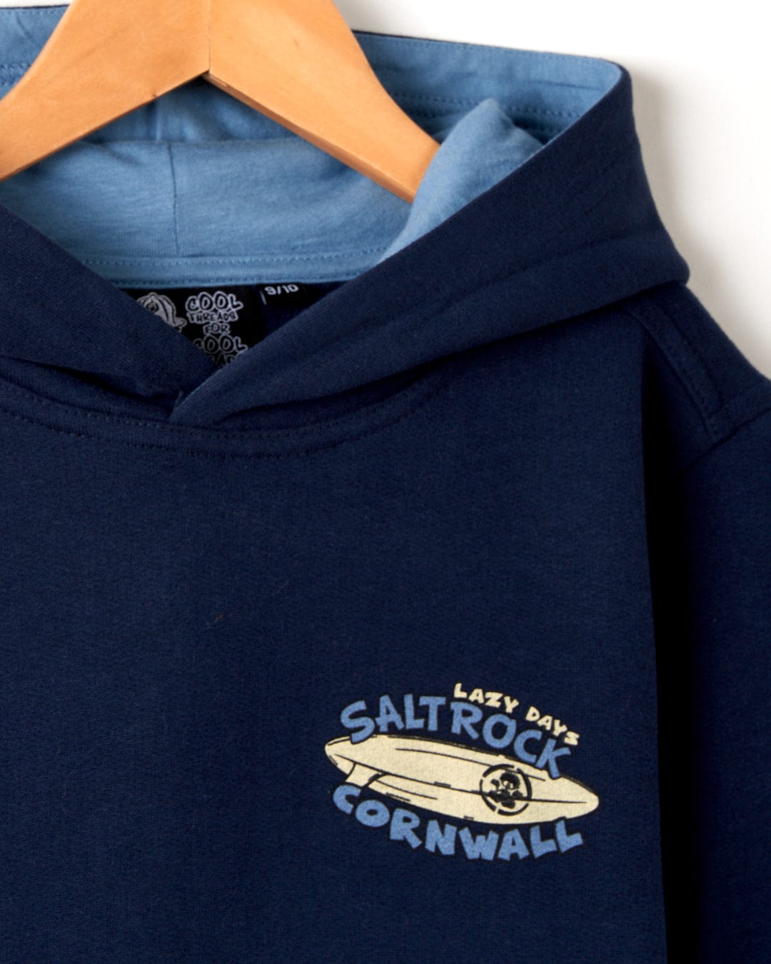 Close-up of a Lazy Location Cornwall - Recycled Kids Hoodie - Blue with the Saltrock logo, overlaid on an orange hanger against a white background.