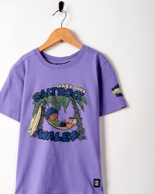 Purple cotton material Lazy Location Wales kids t-shirt with surfing graphics printed on it, hanging on a wooden hanger against a white wall.