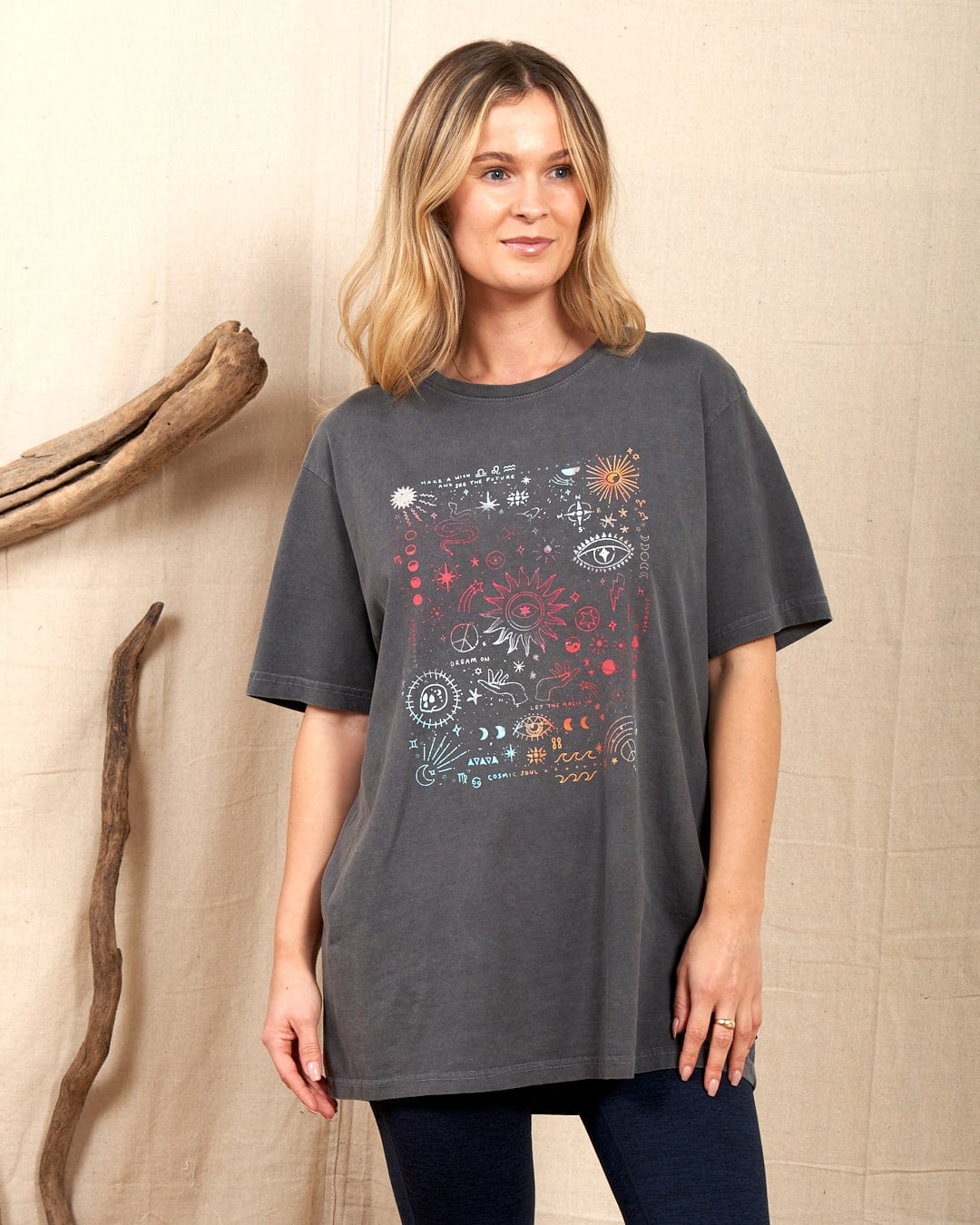 A person models the Saltrock "Let The Magic In" women's t-shirt in washed grey featuring abstract graphics, standing against a neutral background with driftwood.