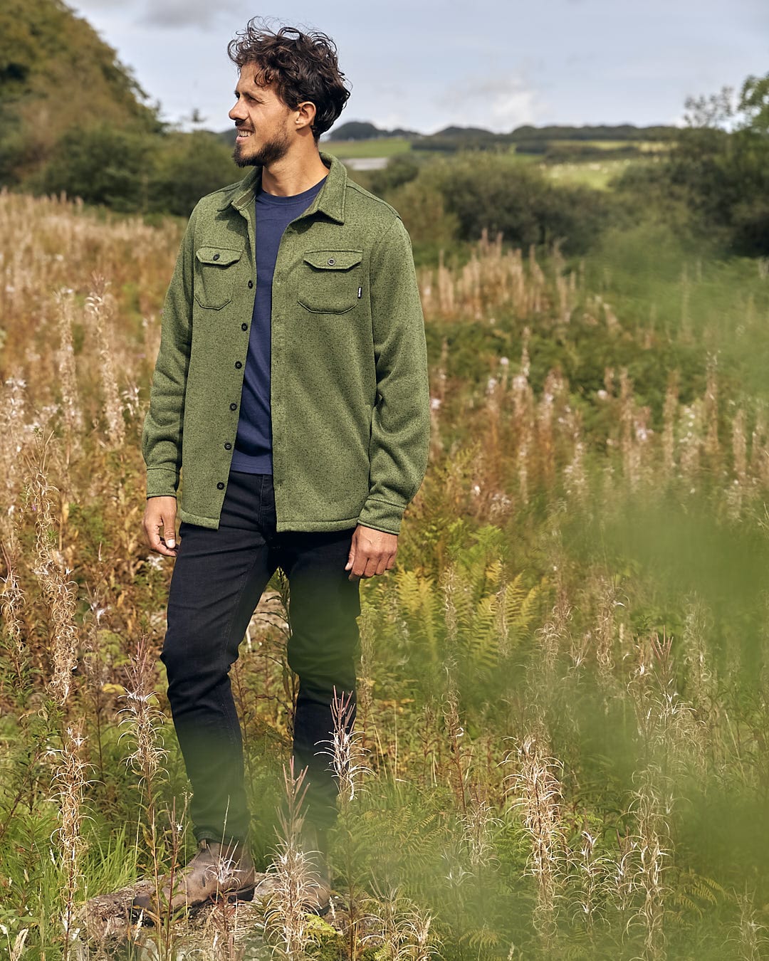 A man with a stylish look wearing a green jacket, featuring the Levick - Men's Long Sleeve Shirt and showcasing Saltrock branding, standing in field.