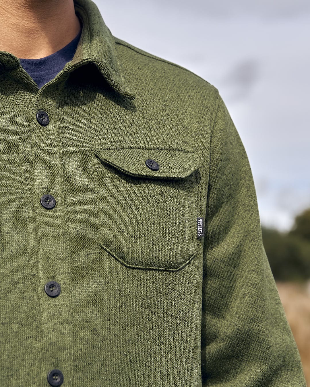 A man in a stylish green Saltrock Levick - Mens Long Sleeve Shirt is standing in a field, representing the brand's fashionable image.