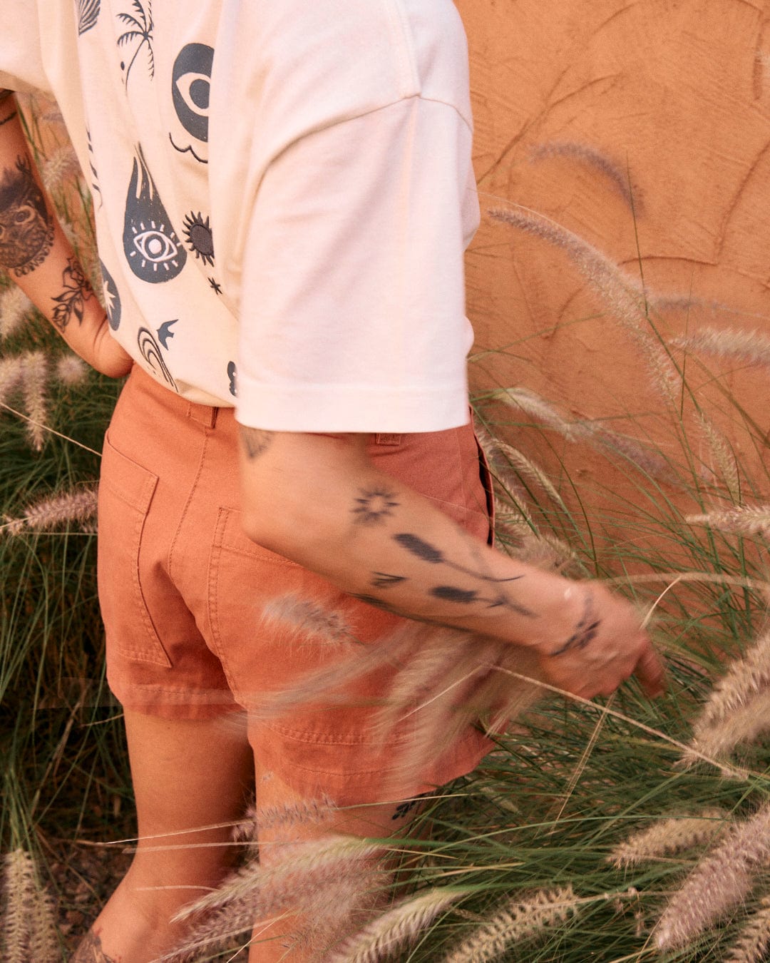 A person with tattoos on their arms wearing a white 100% cotton T-shirt and high-waisted, Liesl - Womens Chino Shorts - Burnt Orange by Saltrock stands among tall grass.