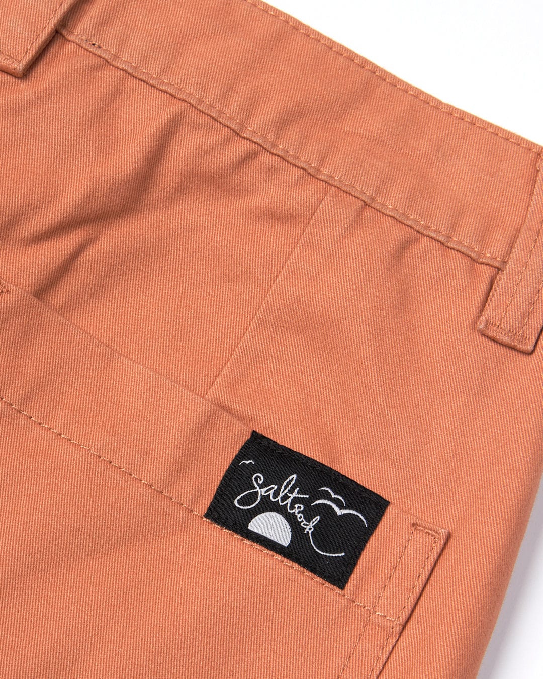 Close-up of a terracotta-colored cotton twill fabric with a black rectangular brand patch labeled "Saltrock" stitched on a pocket.