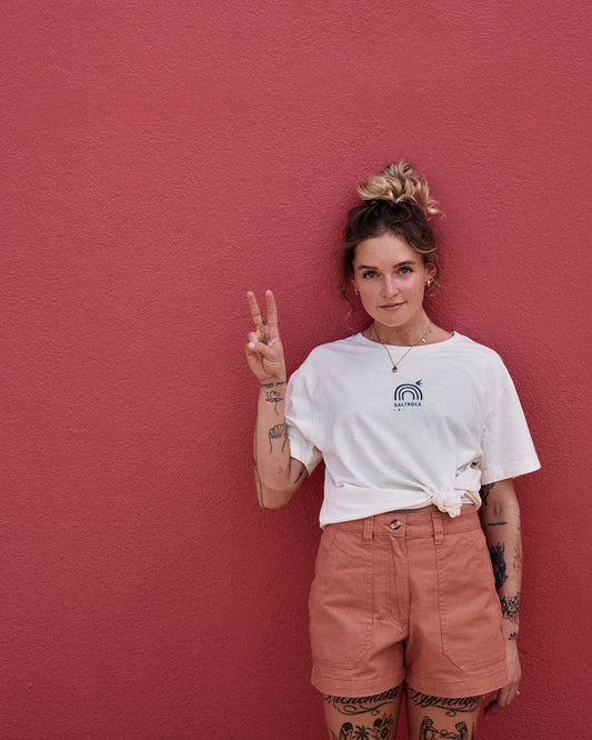A person with tattoos stands against a pink wall, wearing a white T-shirt and Saltrock Liesl - Womens Chino Shorts - Burnt Orange, giving a peace sign with their right hand.