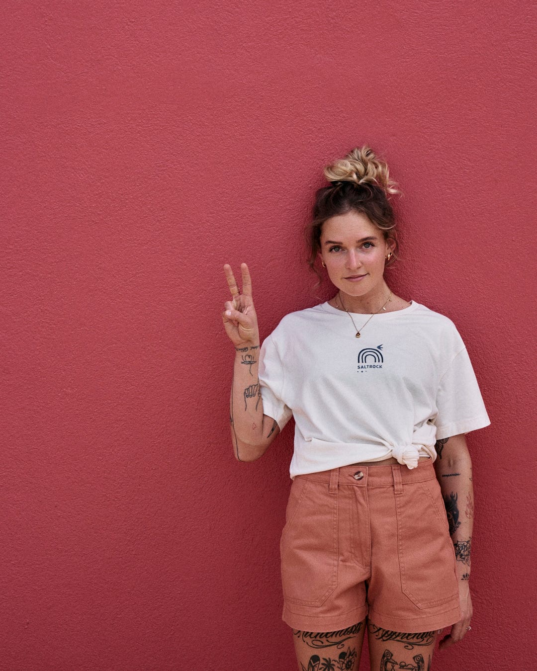 A person with tattoos stands against a pink wall, wearing a white T-shirt and Saltrock Liesl - Womens Chino Shorts - Burnt Orange, giving a peace sign with their right hand.