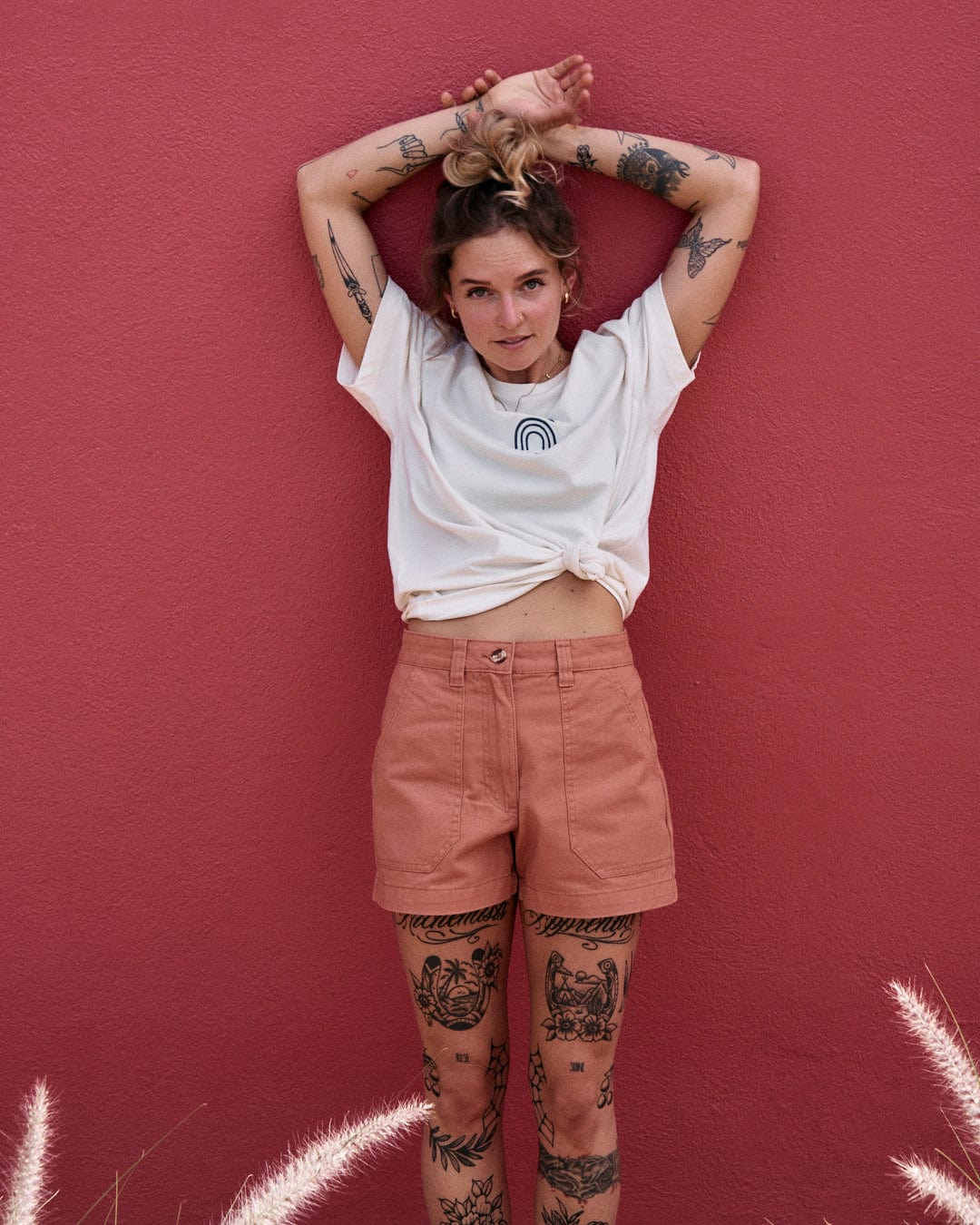 A person with multiple tattoos is standing against a red wall, wearing a 100% cotton white t-shirt and Liesl - Womens Chino Shorts - Burnt Orange by Saltrock, with their hands raised and resting on their head.