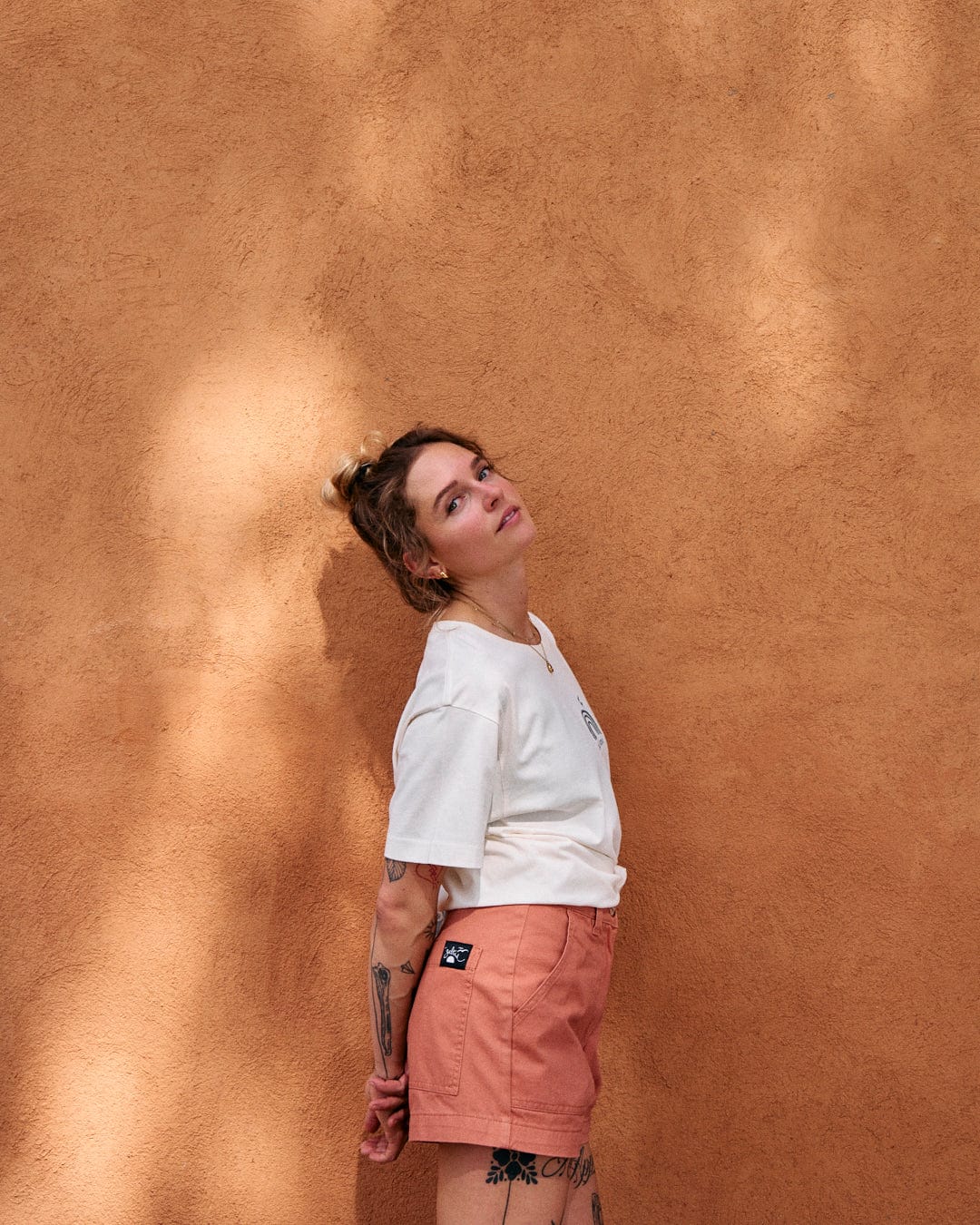 A person with tattoos stands against a burnt orange textured wall, wearing a white shirt and Saltrock's Liesl - Womens Chino Shorts - Burnt Orange, made of 100% cotton, looking upwards.