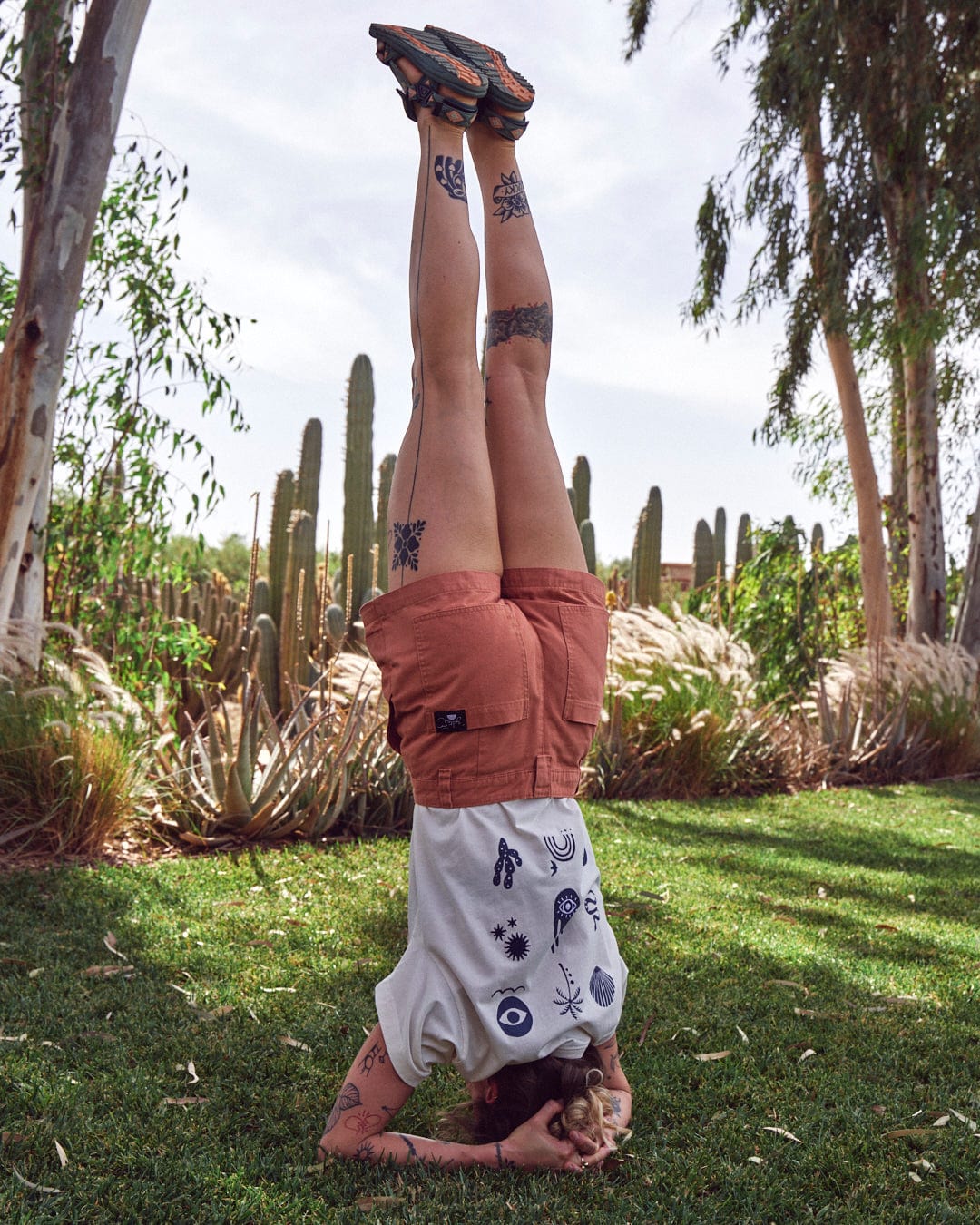 A person in Saltrock Liesl - Womens Chino Shorts - Burnt Orange is performing a headstand on a grassy area surrounded by tall cacti and trees on a sunny day.