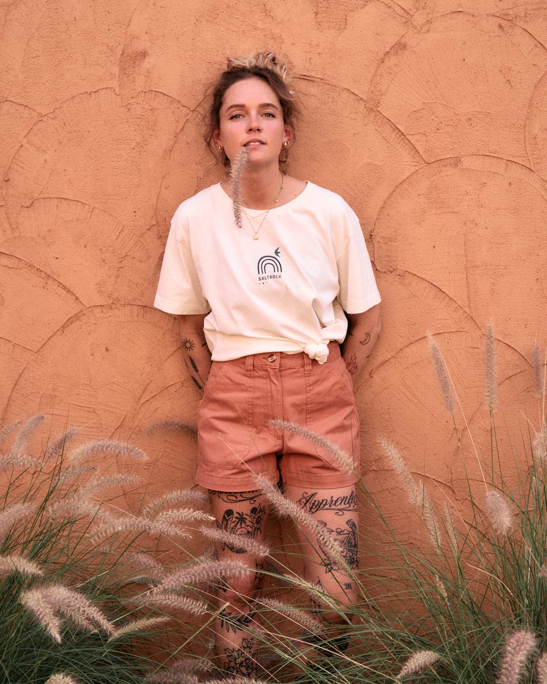 A person with tattoos stands against a textured, burnt orange wall, wearing a white t-shirt made of 100% cotton with a rainbow logo and rolled-up Saltrock Liesl - Womens Chino Shorts - Burnt Orange. Tall grass with white plumes is in the foreground.