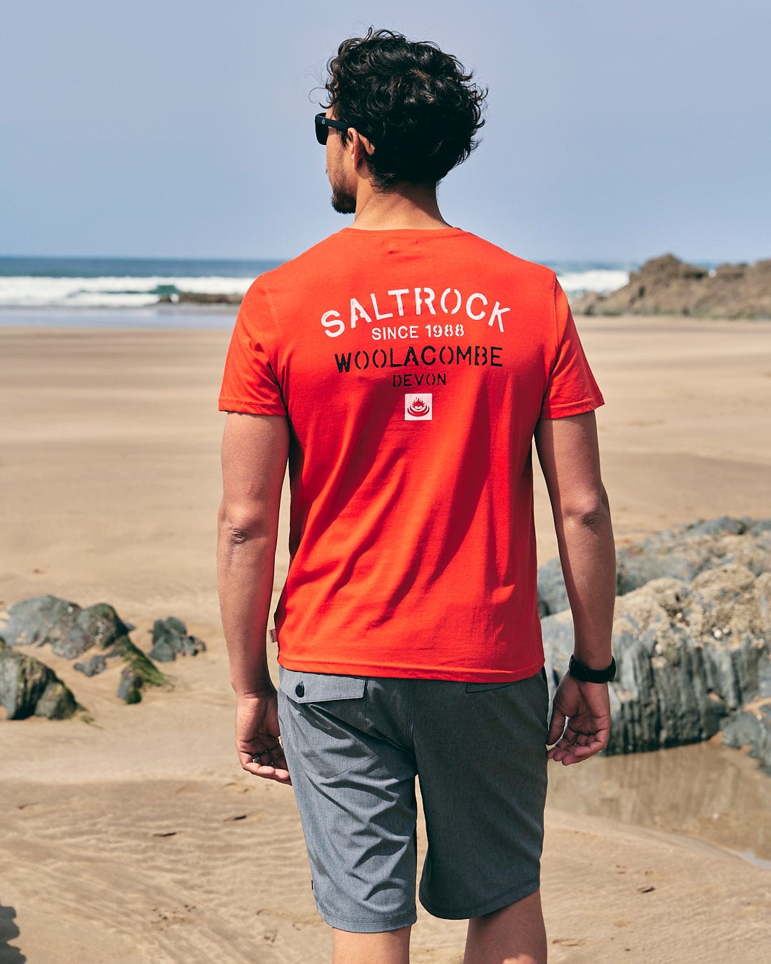 A man walking on the beach wearing a Saltrock Stencil - Mens Location T-Shirt - Woolacombe - Red.