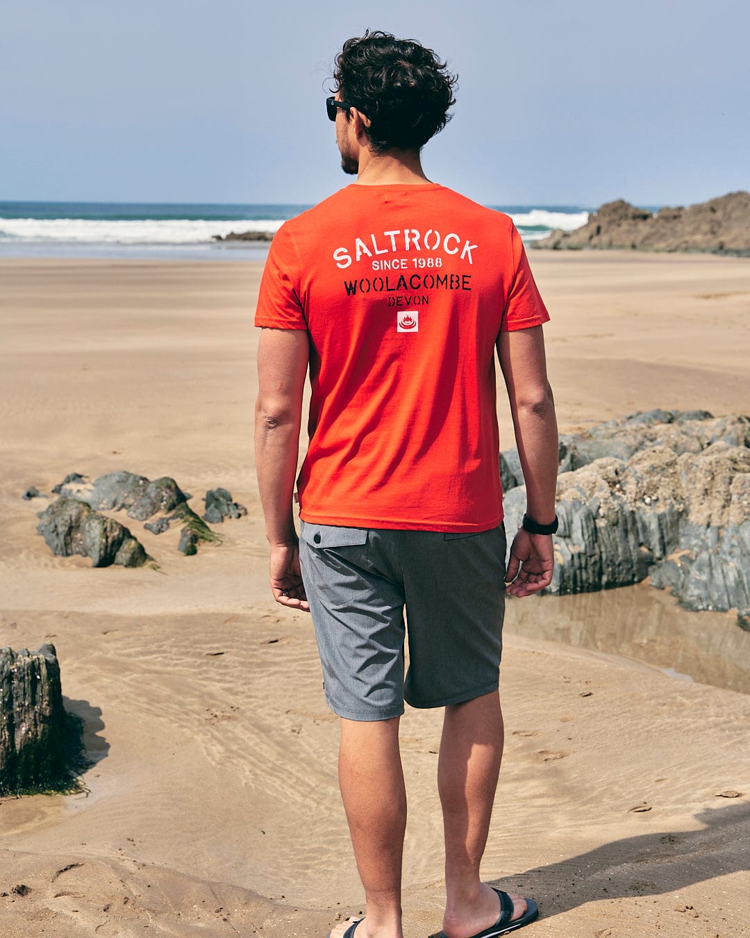 A man wearing a Stencil - Mens Location T-Shirt - Woolacombe - Red by Saltrock standing on a beach.