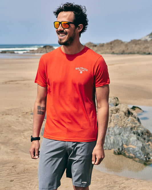 A man wearing sunglasses and a Saltrock Stencil - Mens Location T-Shirt - Woolacombe - Red on the beach.