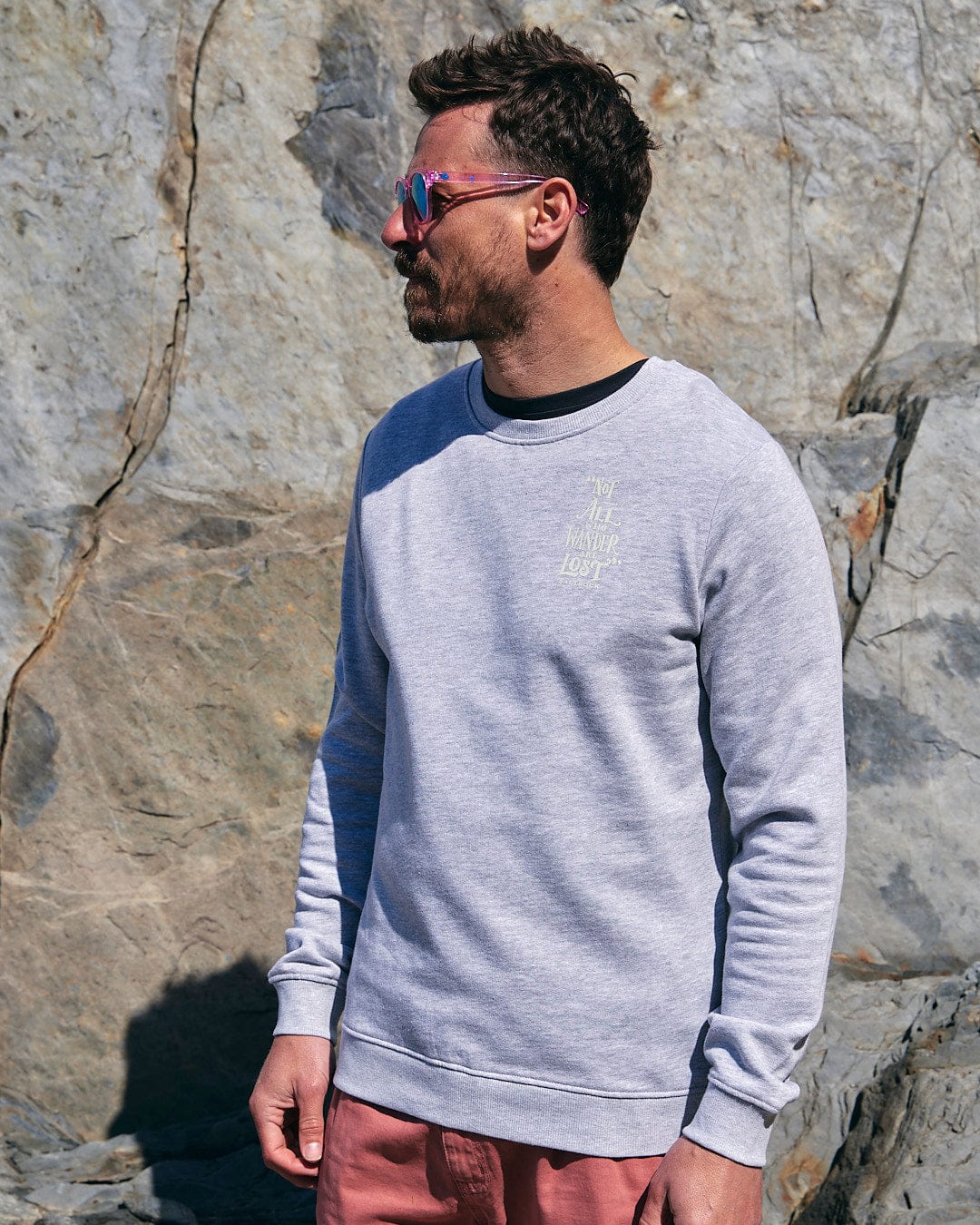 A man wearing a Saltrock Lost Ships - Mens Crew Sweat - Grey sweatshirt and pink shorts standing next to rocks.