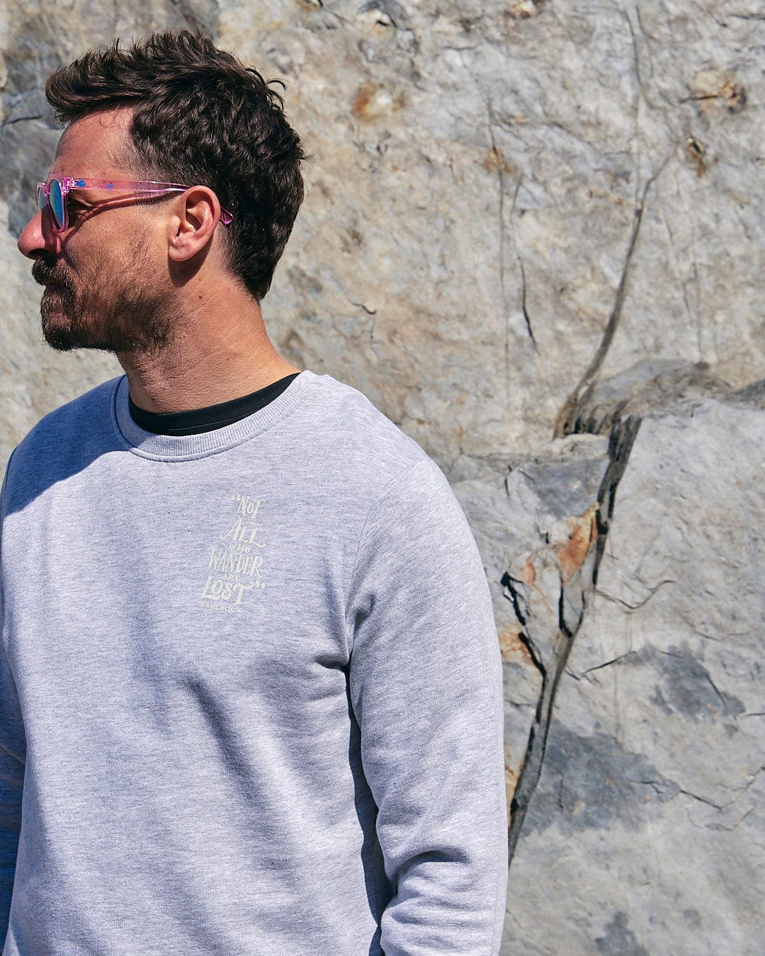 A man wearing Lost Ships - Mens Crew Sweat - Grey by Saltrock, sunglasses, and a grey sweatshirt in front of a rock.