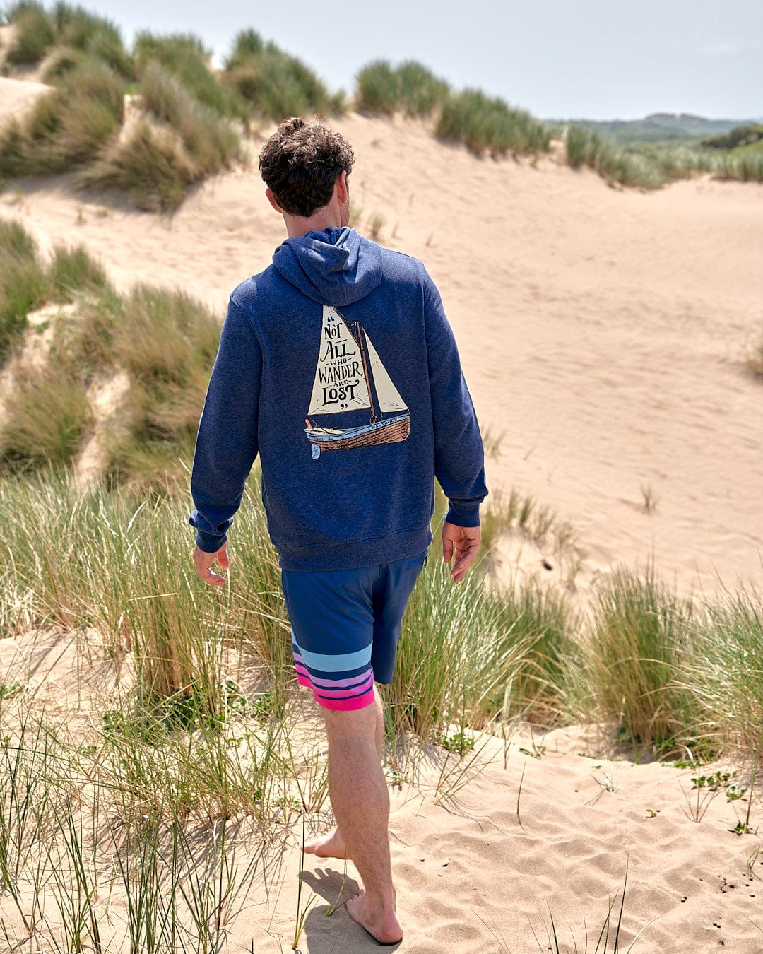 A man wandering through a sand dune with a Lost Ships - Mens Pop Hoodie - Blue Marl from Saltrock on his back.