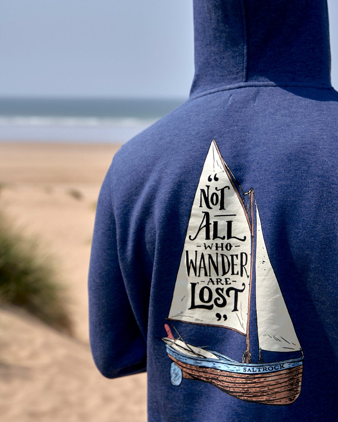 A man wearing a Saltrock Lost Ships - Mens Pop Hoodie in Blue Marl with a sailboat on it and draw cord hood.