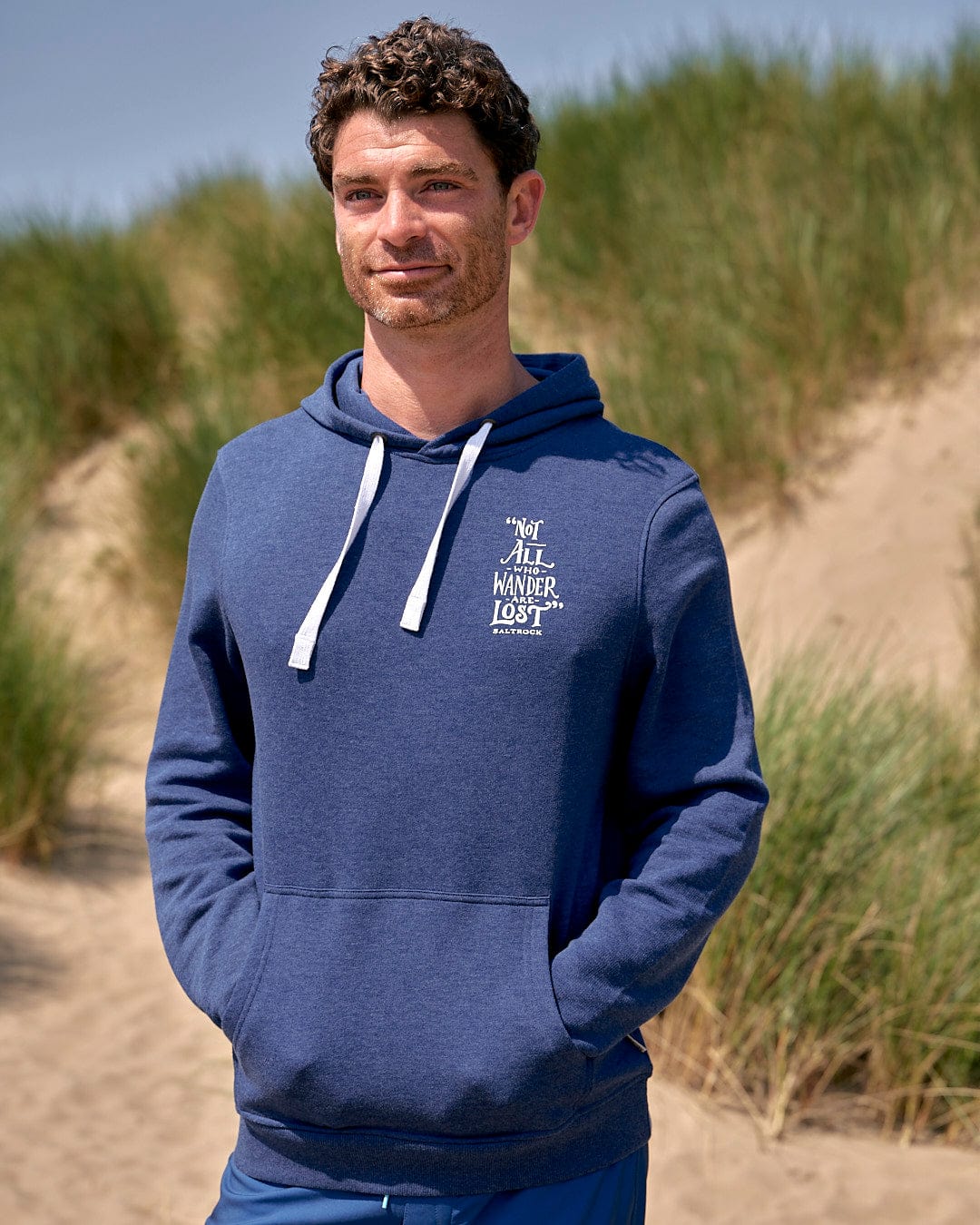 A man standing on a beach wearing a Lost Ships - Mens Pop Hoodie in Blue Marl by Saltrock with a draw cord hood.