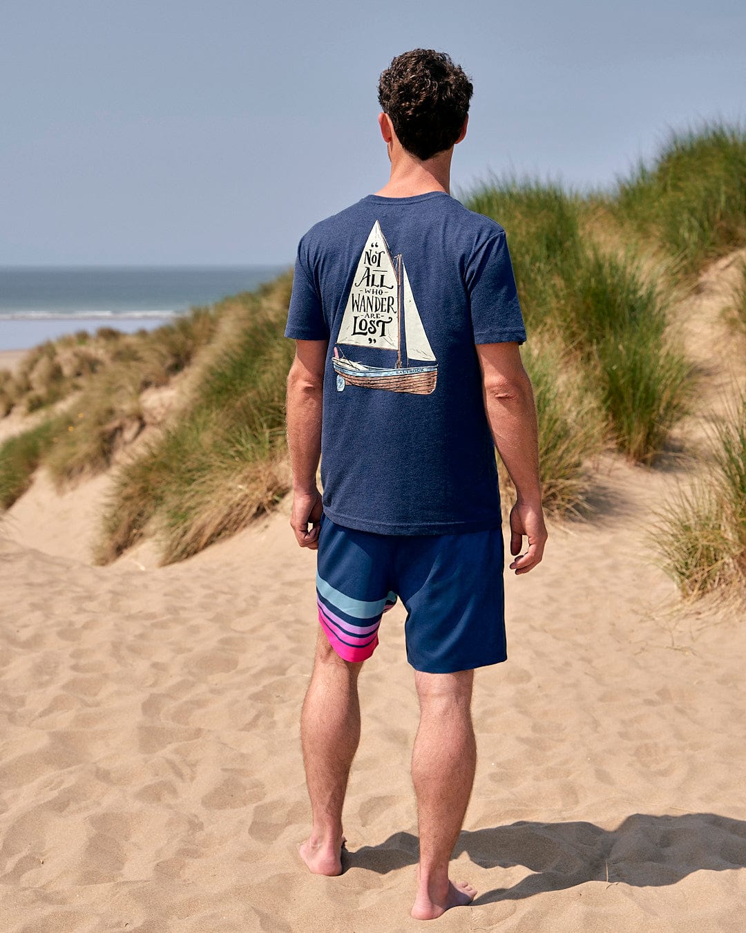 A man standing on a sand dune with a Lost Ships - Mens Short Sleeve T-Shirt - Blue by Saltrock.