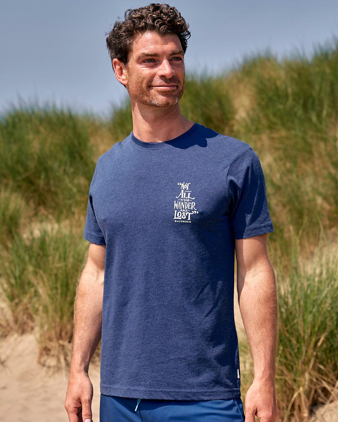 A man standing on a beach wearing a Saltrock Lost Ships - Mens Short Sleeve T-Shirt - Blue.