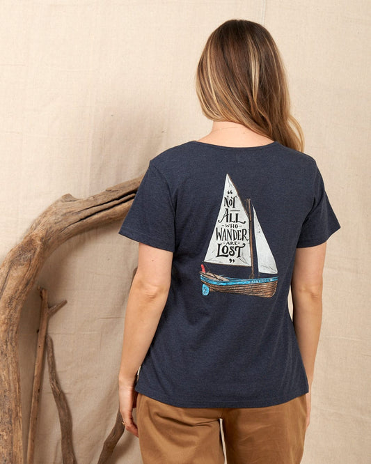 A woman with long hair wears the Saltrock "Lost Ships" women's T-shirt in blue, featuring a sailboat and "Not All Who Wander Are Lost" on the back. She stands before a beige fabric backdrop decorated with driftwood. Made of 100% cotton.