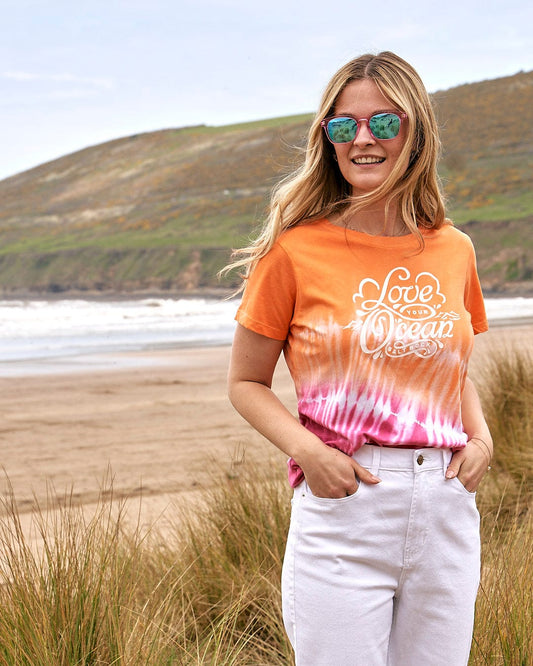 A woman wearing a Love Your Ocean - Tie Dye T-Shirt - Orange from Saltrock standing on the beach while enjoying the ocean.