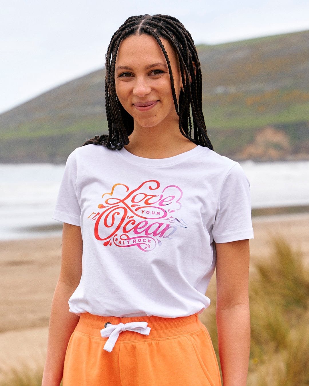 A woman wearing a Saltrock Love Your Ocean - Womens Short Sleeve T-Shirt in White and orange shorts standing on the beach.