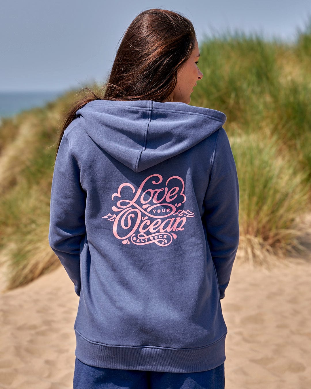 A woman wearing a Saltrock "Love Your Ocean - Womens Zip Hood - Dark Blue" hoodie standing on a beach.