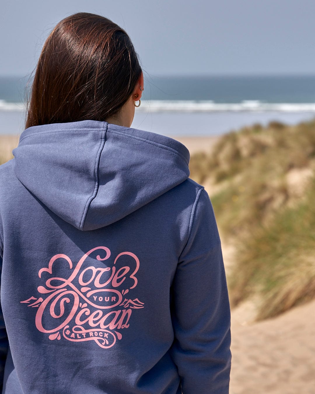 A woman wearing a Saltrock Love Your Ocean - Womens Zip Hood - Dark Blue hoodie standing on the beach.