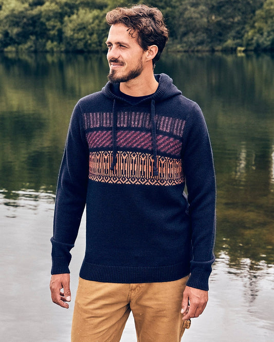 A man wearing a Lukas - Mens Knitted Hoodie from Saltrock stands next to a body of water in the fall and winter season. The hoodie features Saltrock branding.
