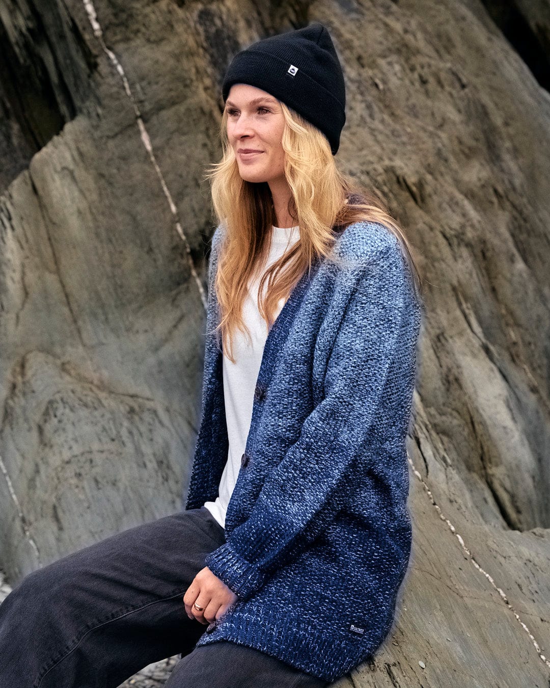 A woman wearing a Saltrock Lynton -  Womens Button Cardigan - Dark Blue sitting on rocks.