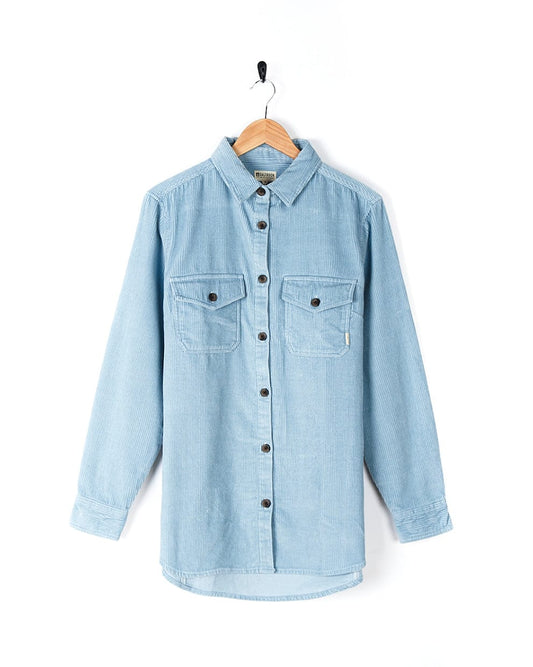 The women's light blue cord Maddox Shirt with buttoned cuffs is hanging on a wooden hanger with Saltrock branding.