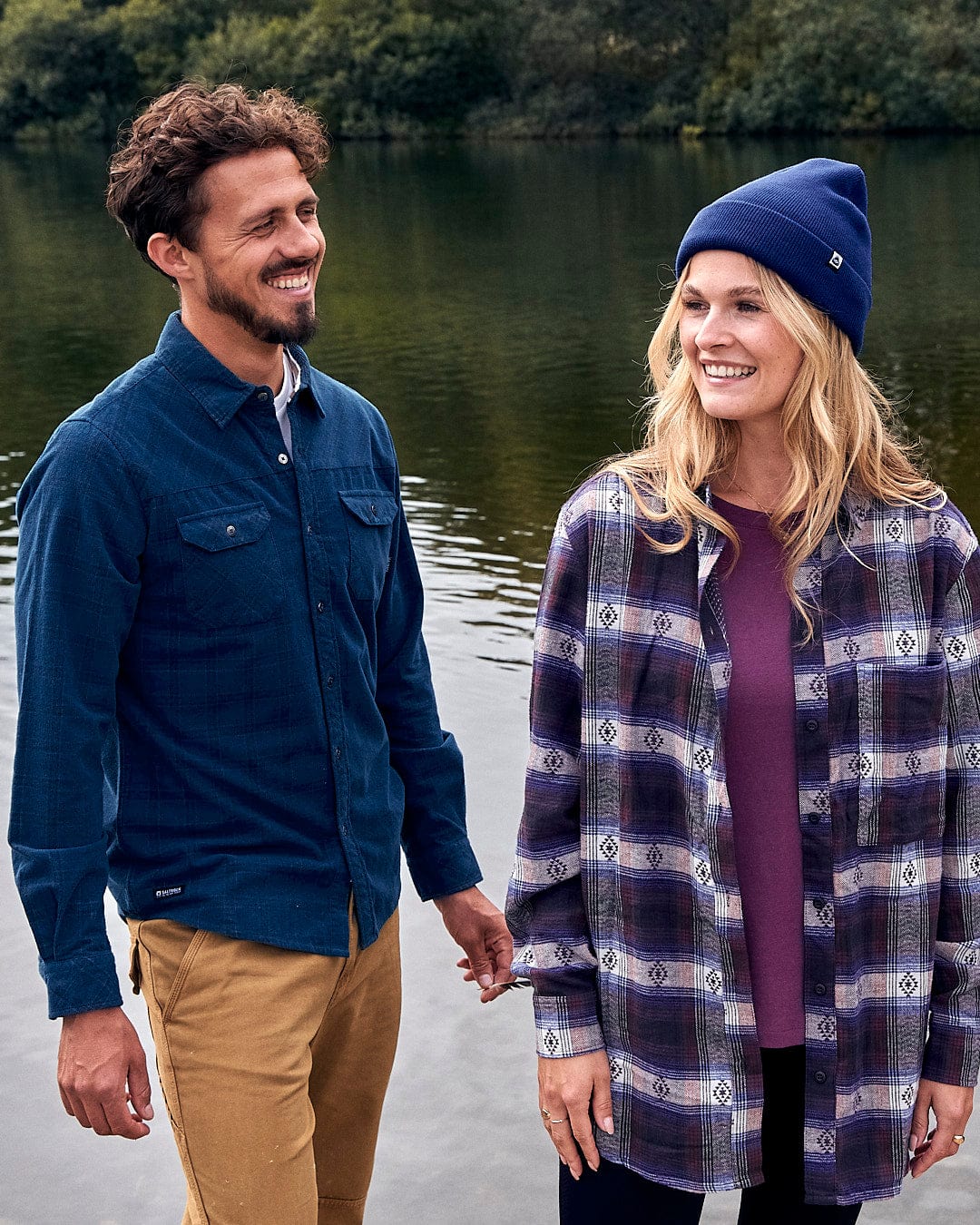 A man and woman standing next to a body of water, wearing the Saltrock Ok - Tight Knit Beanie in Dark Blue.