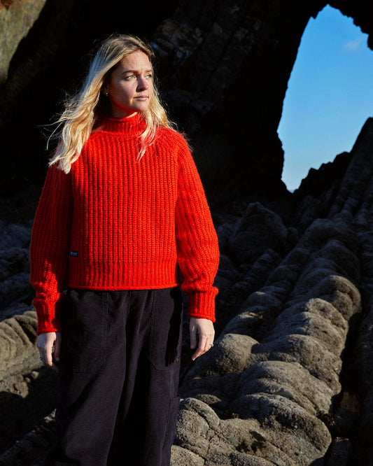 A woman in a Mandy - Womens Funnel Neck Jumper - Red sweater standing in front of a rock formation from Saltrock.