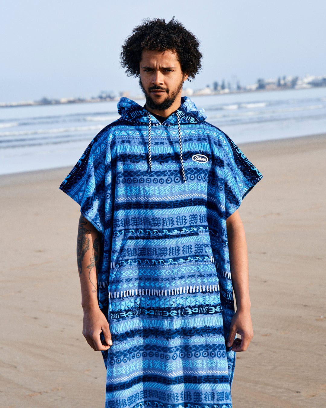 Man with curly hair wearing a Saltrock Changing Towel in Blue with a vintage stripe pattern, standing on a beach with the ocean in the background.