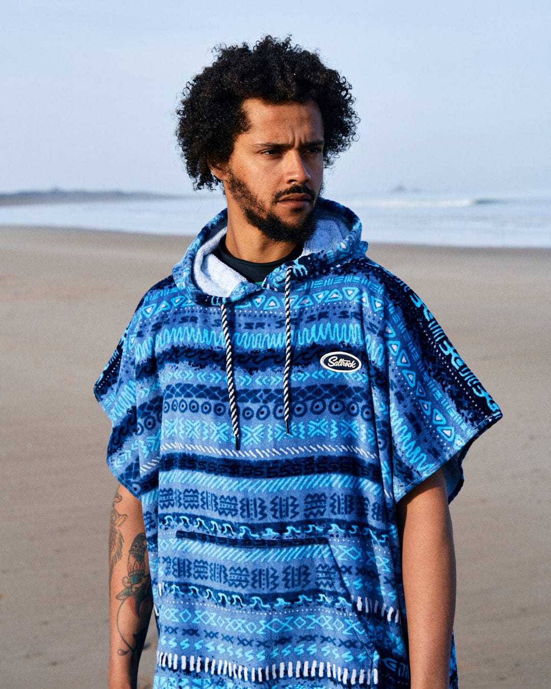 A man with curly hair stands on a beach, wearing a Saltrock blue hoodie with a vintage stripe pattern, looking to the side.