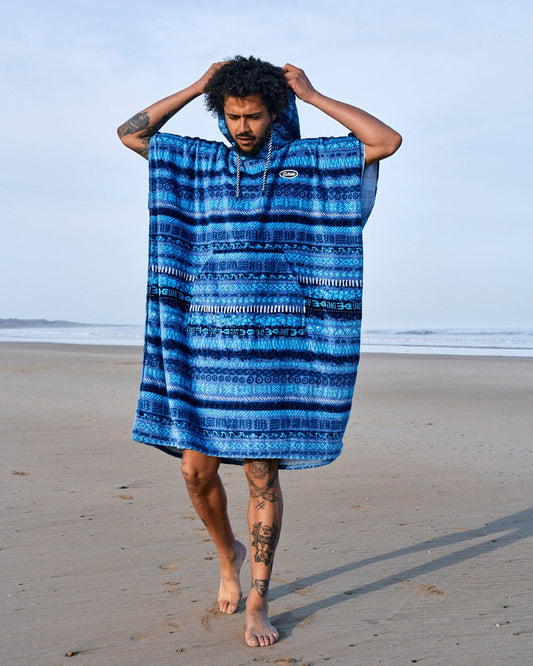 Man with tattoos wearing a blue Saltrock Changing Towel, standing barefoot on a sandy beach, adjusting the hood over his head.
