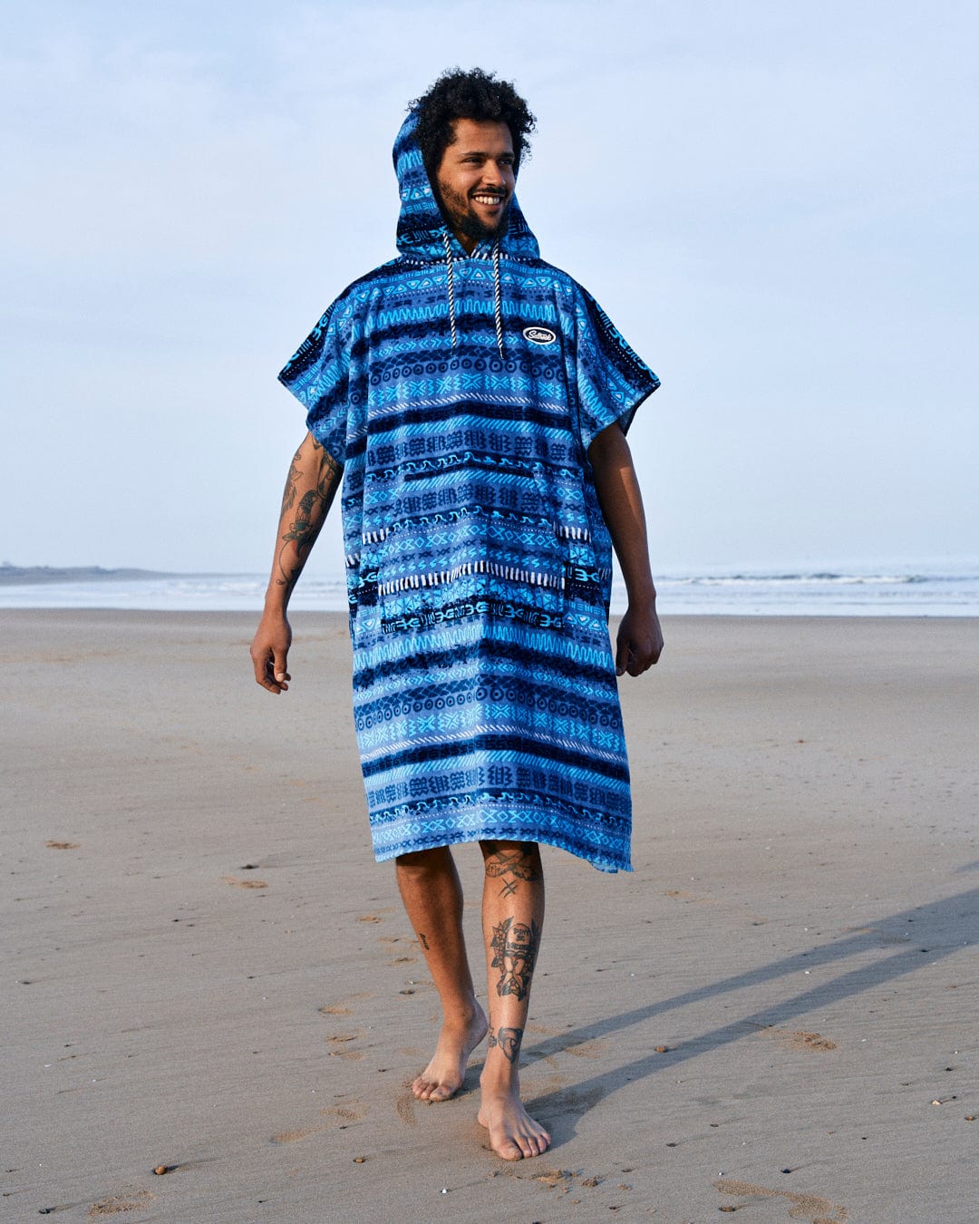 A man with a beard and tattoos smiles while wearing a Saltrock Marks Changing Towel in Blue on a sandy beach.