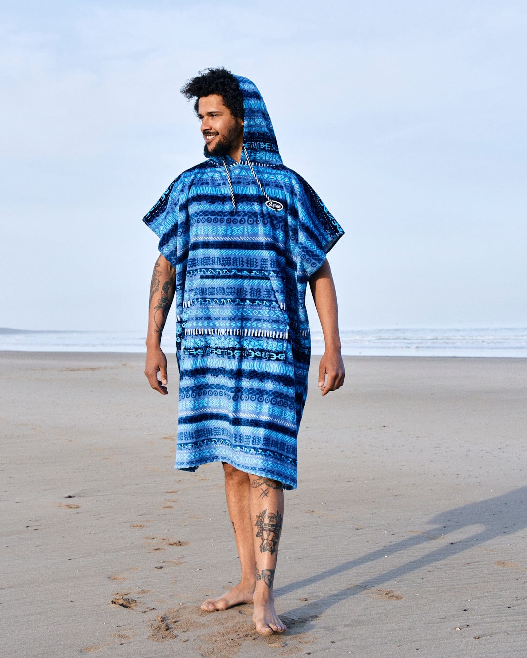 A man in a Saltrock blue vintage stripe pattern Marks - Changing Towel poncho standing barefoot on a sandy beach, looking to the side with a slight smile.