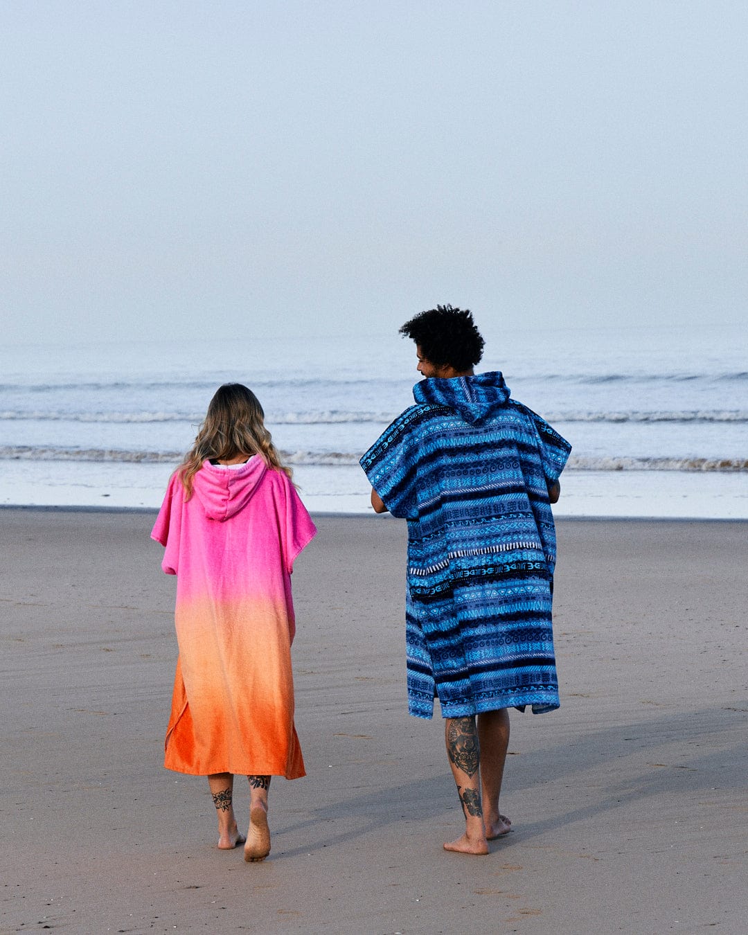 Two people in colorful Saltrock Marks - Changing Towel - Blue towels walking barefoot on a beach, viewed from behind, with the ocean in the background.