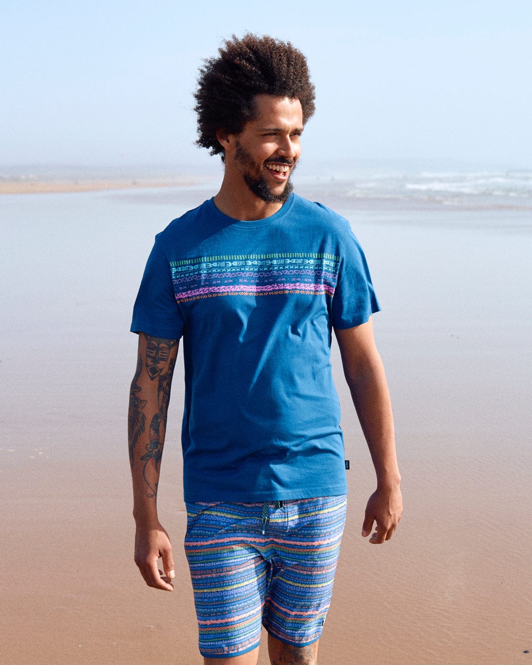 A man with curly hair and a tattoo on his arm wearing a Saltrock Marks Chest - Mens Short Sleeve T-Shirt in Blue and shorts standing on a beach.