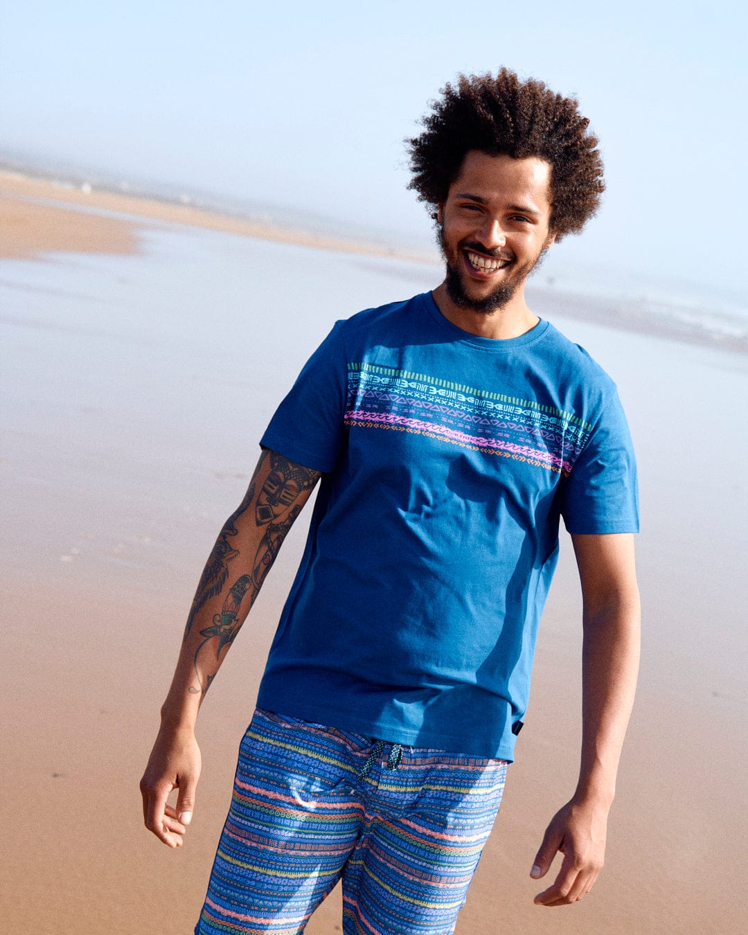 A young man with curly hair smiling, wearing a Marks Chest - Mens Short Sleeve T-Shirt in Blue from Saltrock, and patterned shorts, walking on a sandy beach.