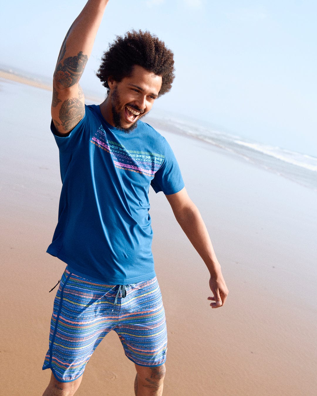 A joyful man with a beard and tattoos raises his arm while walking on a sandy beach, wearing a Marks Chest - Mens Short Sleeve T-Shirt in Blue made of peached soft material and colorful shorts.