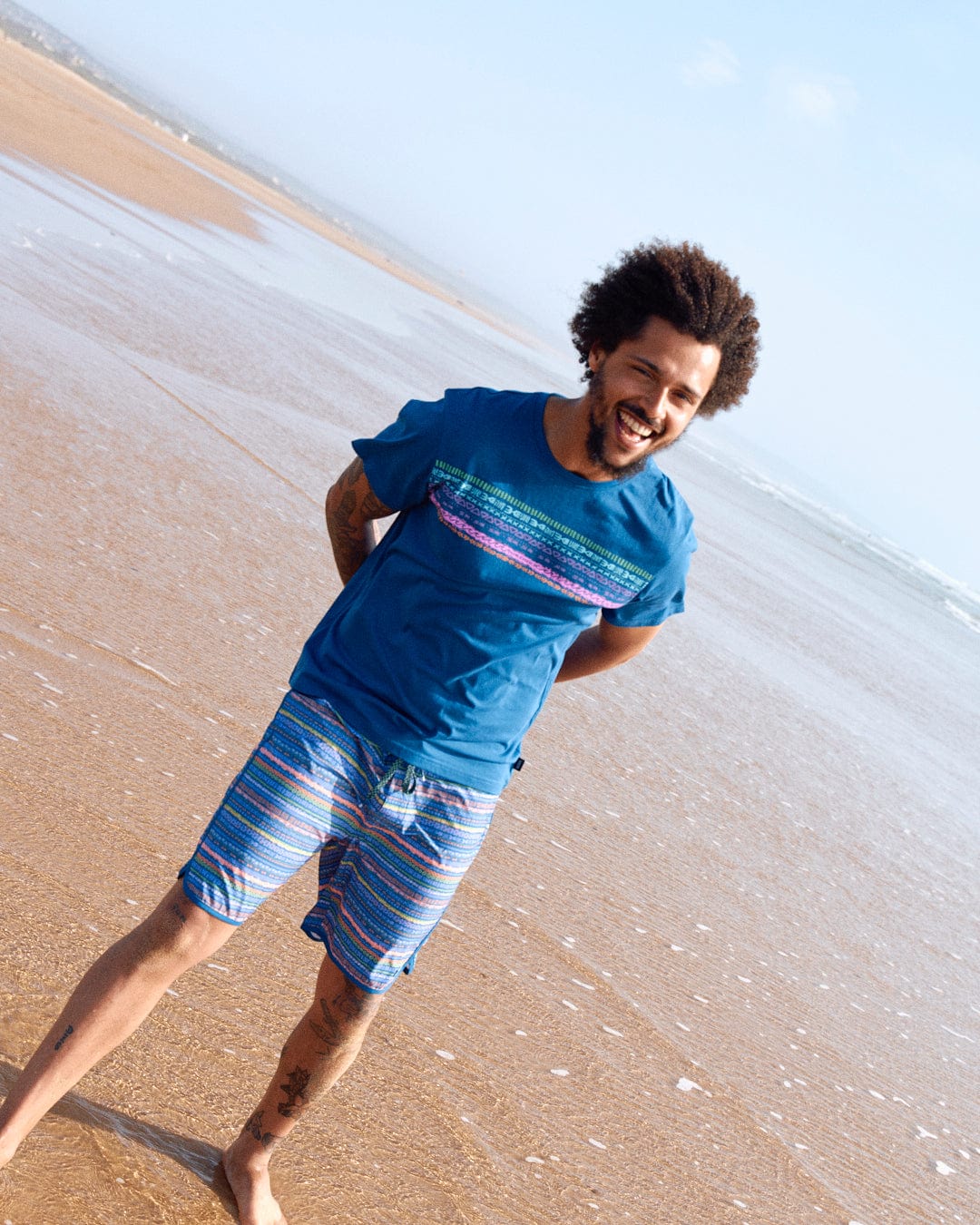 Man with curly hair and tattoos smiles while walking on a beach, wearing a Saltrock Marks Chest Mens Short Sleeve T-Shirt in Blue and shorts.