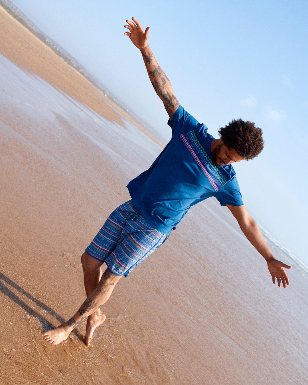 A young man with a tattoo on his arm balances on one leg on a sandy beach, wearing a Marks Chest - Mens Short Sleeve T-Shirt in Blue by Saltrock, and patterned shorts, his arms outstretched.
