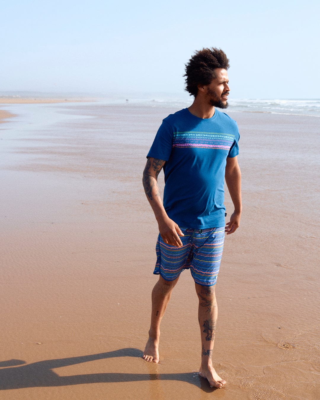 A man in a Saltrock Marks Chest - Mens Short Sleeve T-Shirt in Blue with crew neckline and striped shorts standing on a sandy beach, looking to the side, with ocean waves in the background.